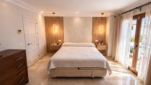 Une chambre avec un grand lit, une tête de lit en bois, des tables de chevet, une commode et une porte-fenêtre coulissante donnant sur un balcon.