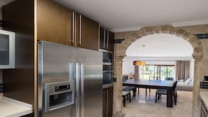 Une cuisine moderne équipée d’un réfrigérateur en acier inoxydable, d’un four encastré et d’un four à micro-ondes. On y trouve un coin repas avec une table et des chaises, ainsi qu’une structure en pierre en forme d’arche qui mène à un espace détente offrant une vue sur l’extérieur.