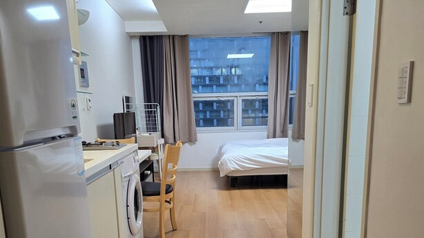 Habitación de hotel compacta con kitchenette, lavadora, cama y vista del paisaje urbano a través de la ventana.