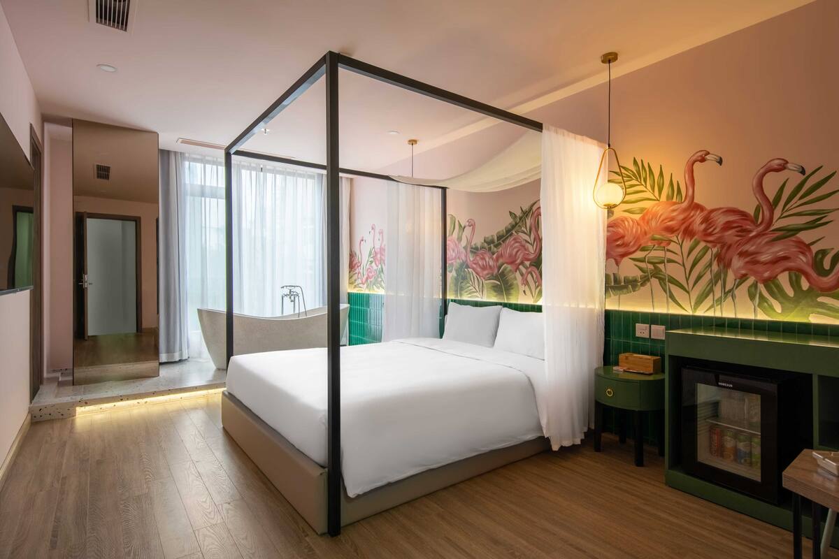 Une chambre d’hôtel moderne avec un lit à baldaquin, un mur d’accent vert orné d’une fresque représentant un flamant rose, un petit coin salon avec une table et un mini-réfrigérateur intégré.
