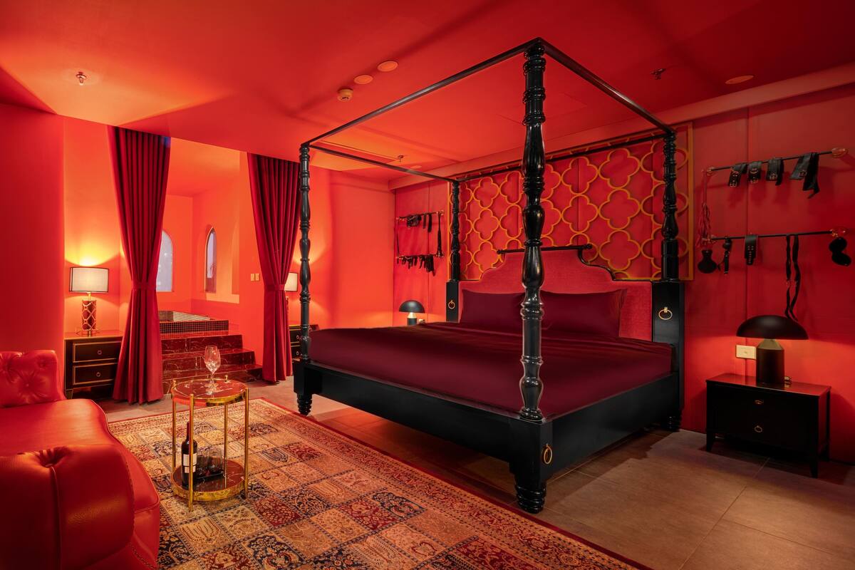 Un lit à baldaquin rouge, une petite table ronde avec un plateau en verre et un canapé rouge dans une pièce aux murs rouges et ornée d’un tapis à motifs.