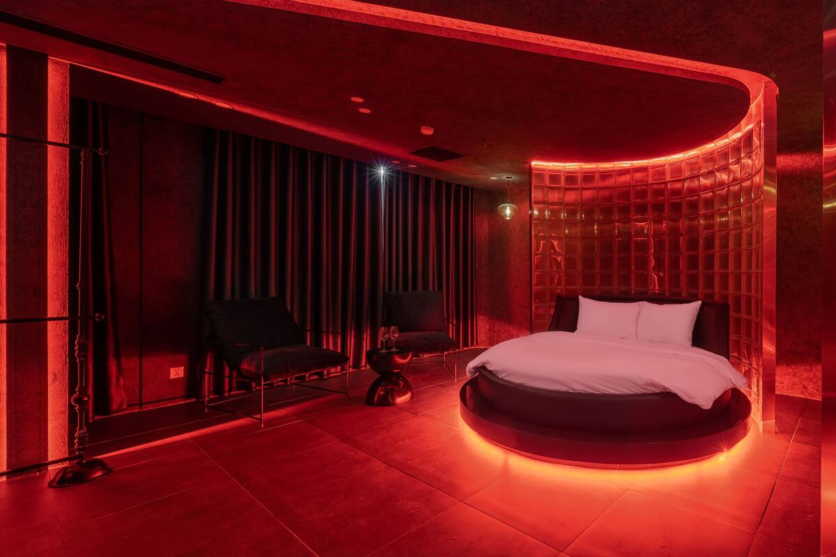 Une chambre à coucher moderne avec un lit aux lignes courbes, un coin salon comprenant un fauteuil et une petite table, ainsi qu’une grande fenêtre agrémentée de rideaux.