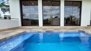 Ein Schwimmbecken mit gefliestem Sonnendeck, großen Schiebeglastüren und Blick auf die Umgebung.