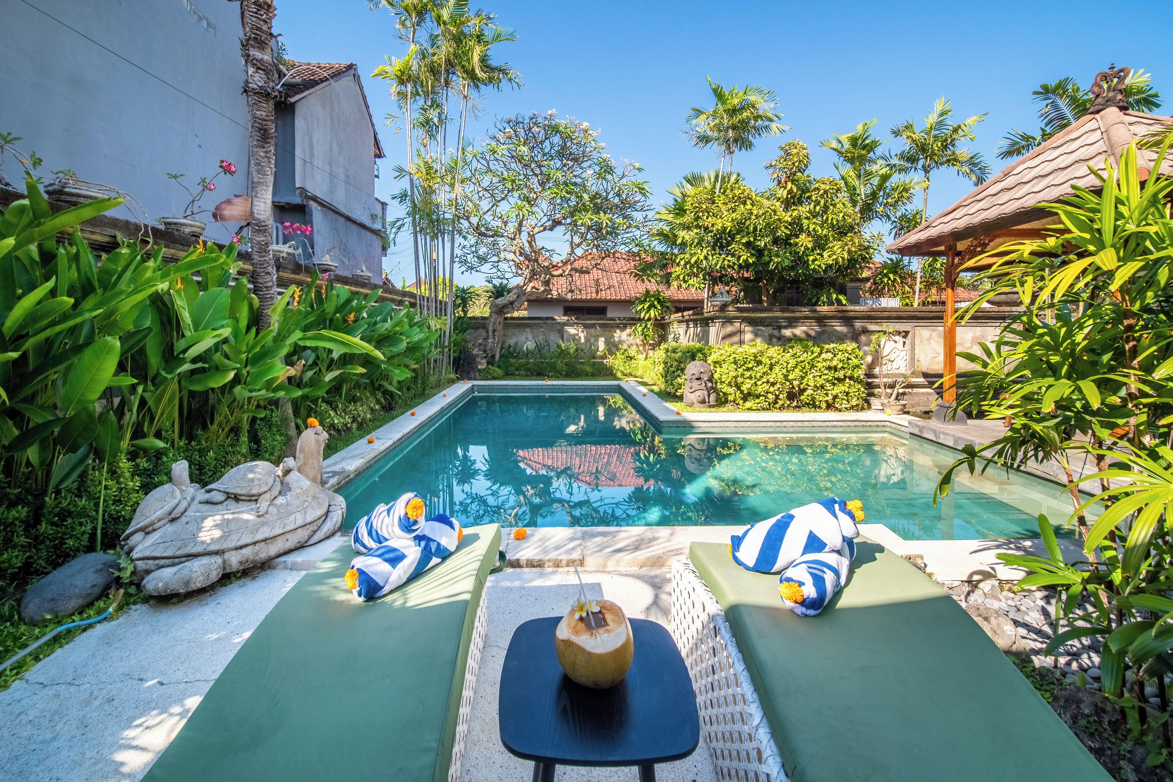 A swimming pool with two lounge chairs and a table, surrounded by tropical plants and a stone statue.