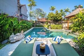 Une piscine bordée de deux chaises longues et d’une table, le tout entouré de plantes tropicales et d’une statue en pierre.
