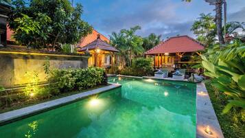 Une piscine dotée d’une terrasse carrelée, entourée de plantes tropicales et bordée de deux petits bâtiments aux toits rouges.