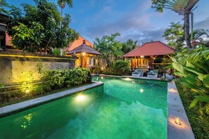 A swimming pool with a tiled deck, surrounded by tropical plants and two small structures with red roofs. - Villa Hibiscus Sanur by Nagisa Bali (Denpasar)