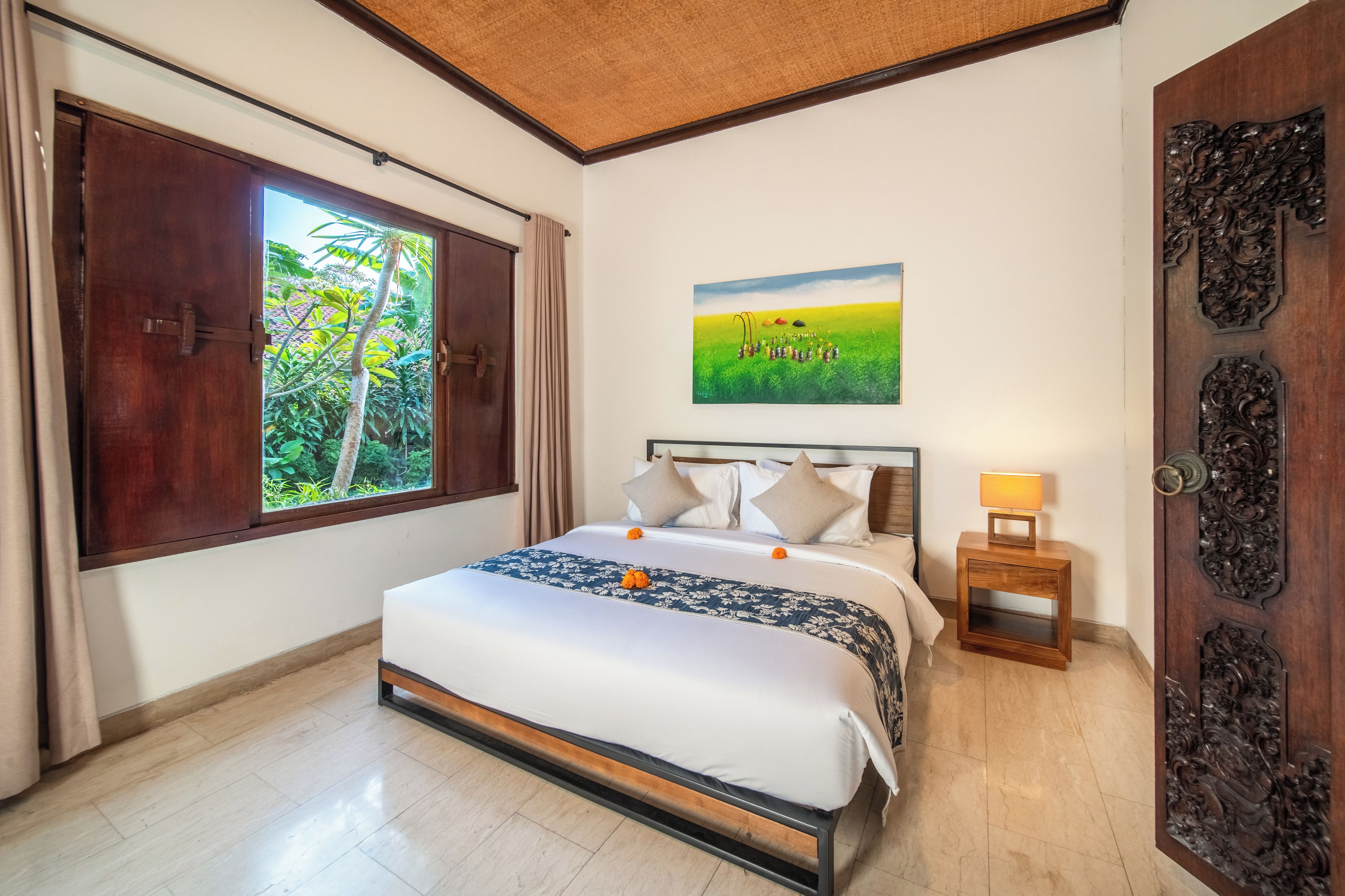 A bedroom with a bed, a nightstand, a window with curtains, a door with intricate carvings, and a painting on the wall.
