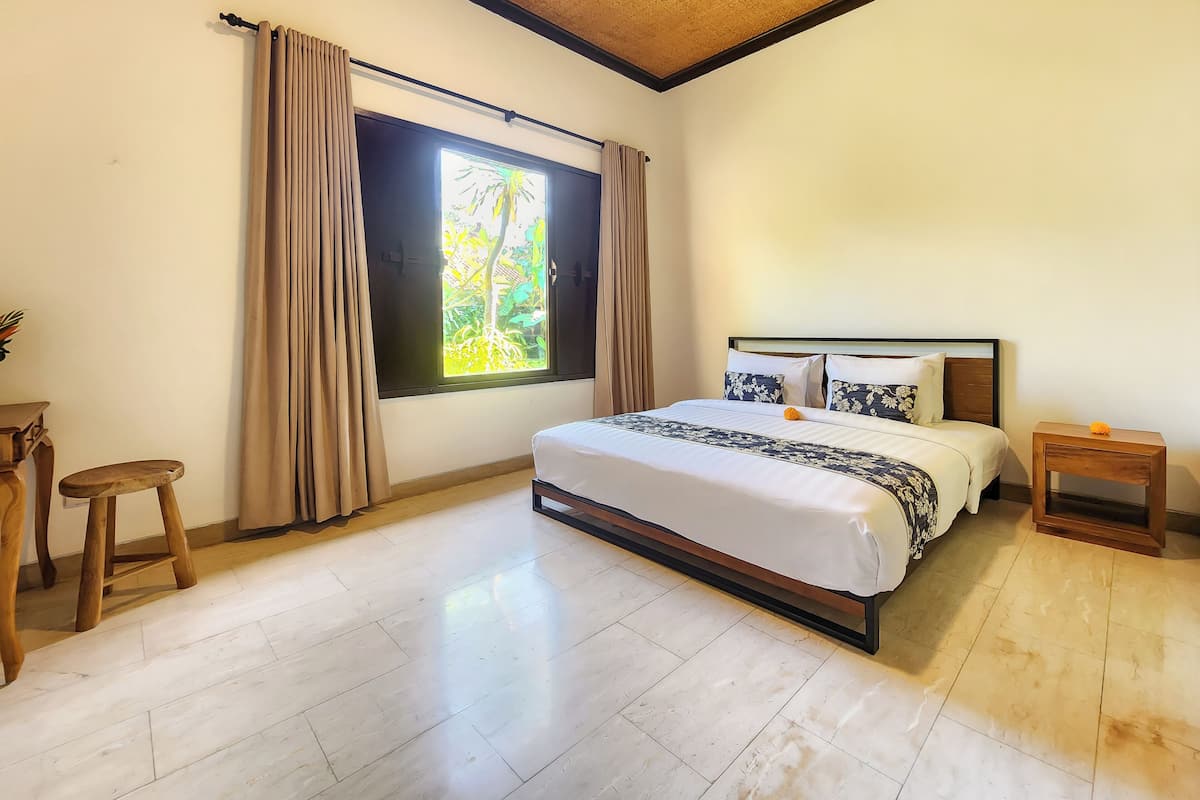 A bedroom with a bed, a window with curtains, a wooden stool, and a nightstand.