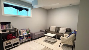 A living room with a grey sofa, a white coffee table, a bookshelf, and a window with blinds.