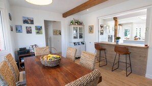 Un coin repas avec une table en bois, des chaises en osier, un panier de fruits, un meuble contenant des bouteilles et une cuisine visible à travers une porte ouverte.