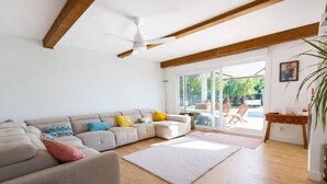 Un salon moderne avec un canapé d’angle, un ventilateur de plafond, un plancher en bois et une vue sur une terrasse extérieure, accessible par des portes vitrées.