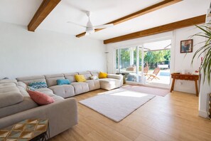 Ein modernes Wohnzimmer mit einem Ecksofa, einem Deckenventilator, Holzboden und Blick durch Glastüren auf eine Terrass.