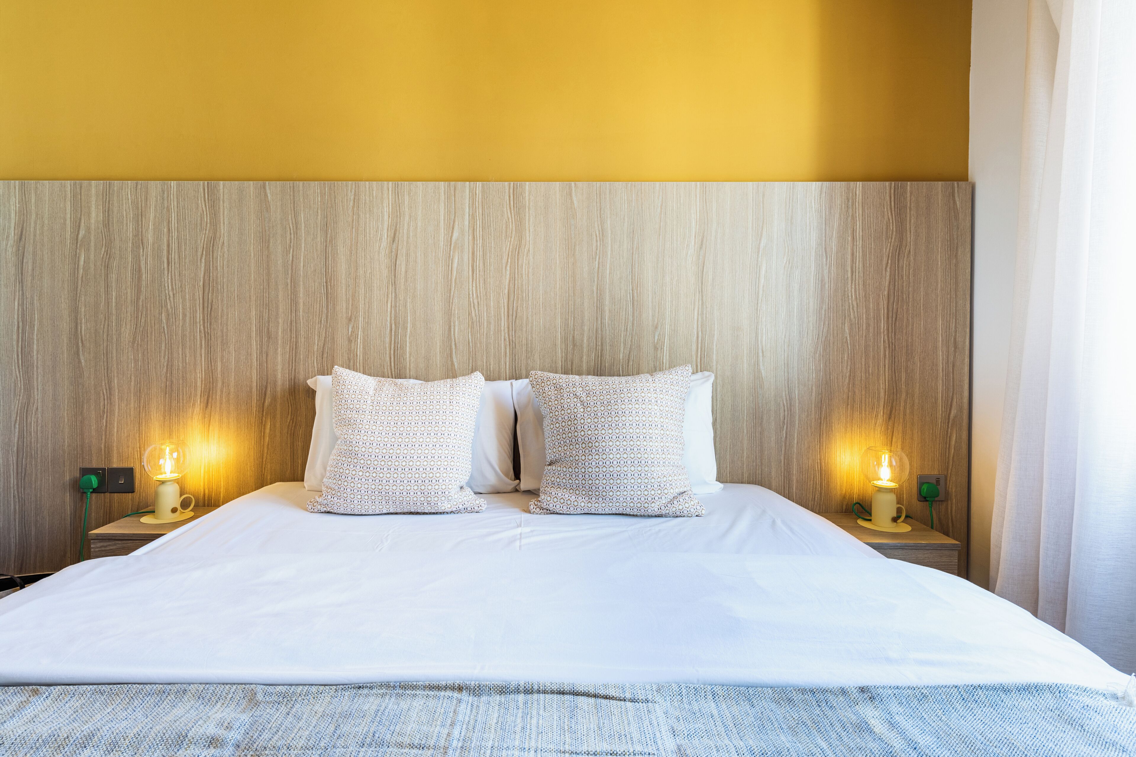 Una cama bien tendida con sábanas blancas y almohadas estampadas, cabecera de madera y lámparas de noche con cables verdes.