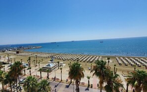 A beach with rows of umbrellas and palm trees, a marina with boats, and a clear blue sky. - Made with love 1-BR By Finikoudes Beach (Larnaca)