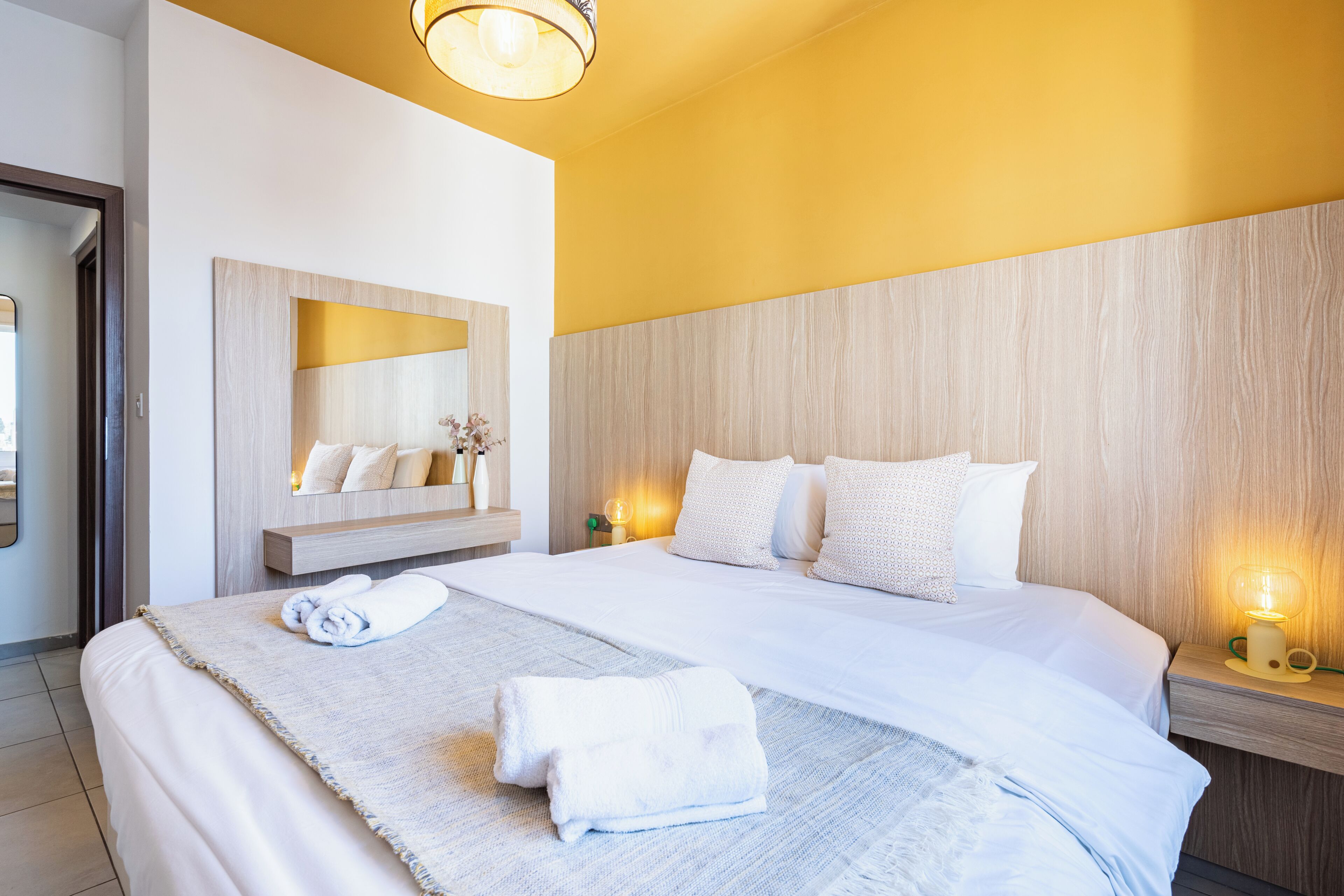 A hotel room with a large bed, a bedside table with a lamp, a mirror, and a wall-mounted shelf with toiletries.
