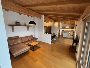 Ein modernes Wohnzimmer mit einer Couch, einem Couchtisch und einer Pendelleuchte. Im Hintergrund sind eine Küche und ein Essbereich zu sehen.
