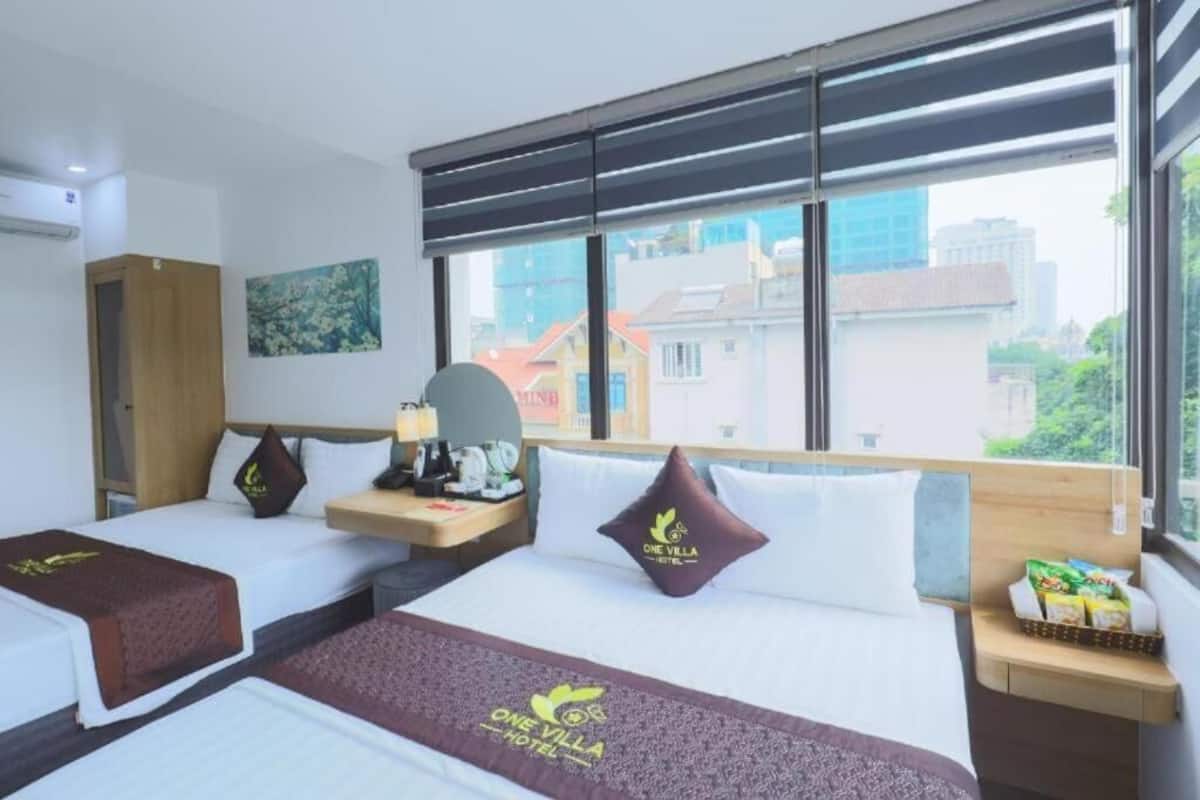 A hotel room with two beds, a wooden headboard, a nightstand, a window with blinds, and a view of buildings outside.
