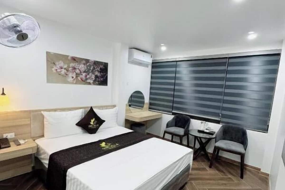 A modern hotel room with a bed, bedside table, desk, chair, and a large window with blinds.