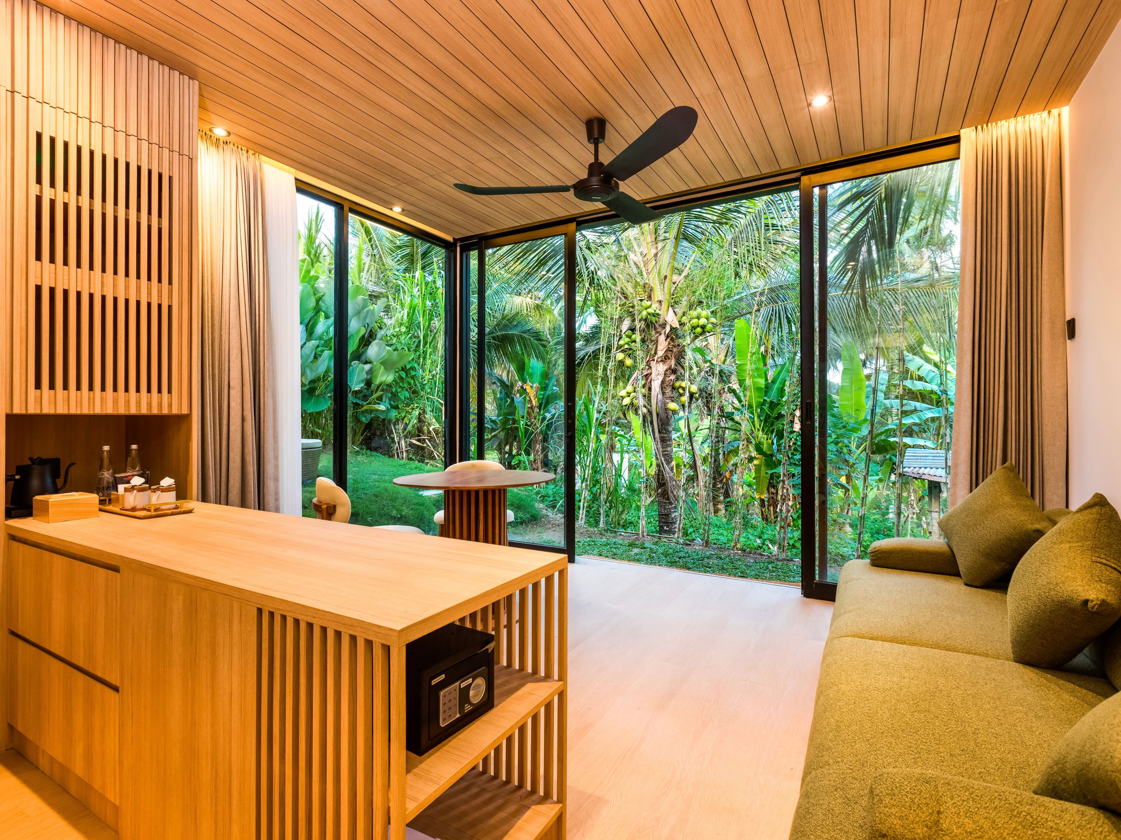 Un interior moderno con techo de madera, ventilador de techo, zona de bar de madera, sofá y vista a exuberante follaje exterior.
