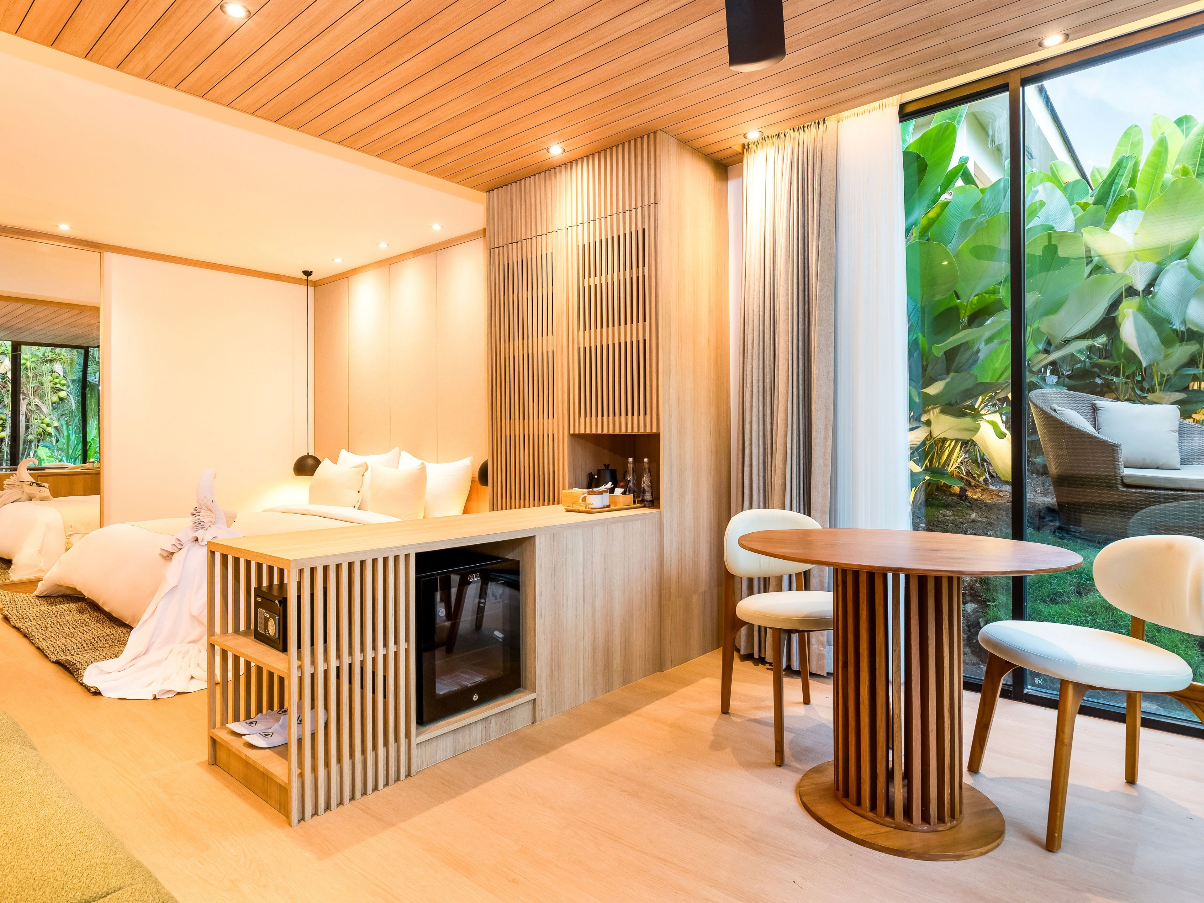 Una habitación de hotel moderna con techo de madera, un ventanal con cortinas, cama con sábanas blancas, una mesita de noche de madera y una mesa de comedor redonda con sillas.