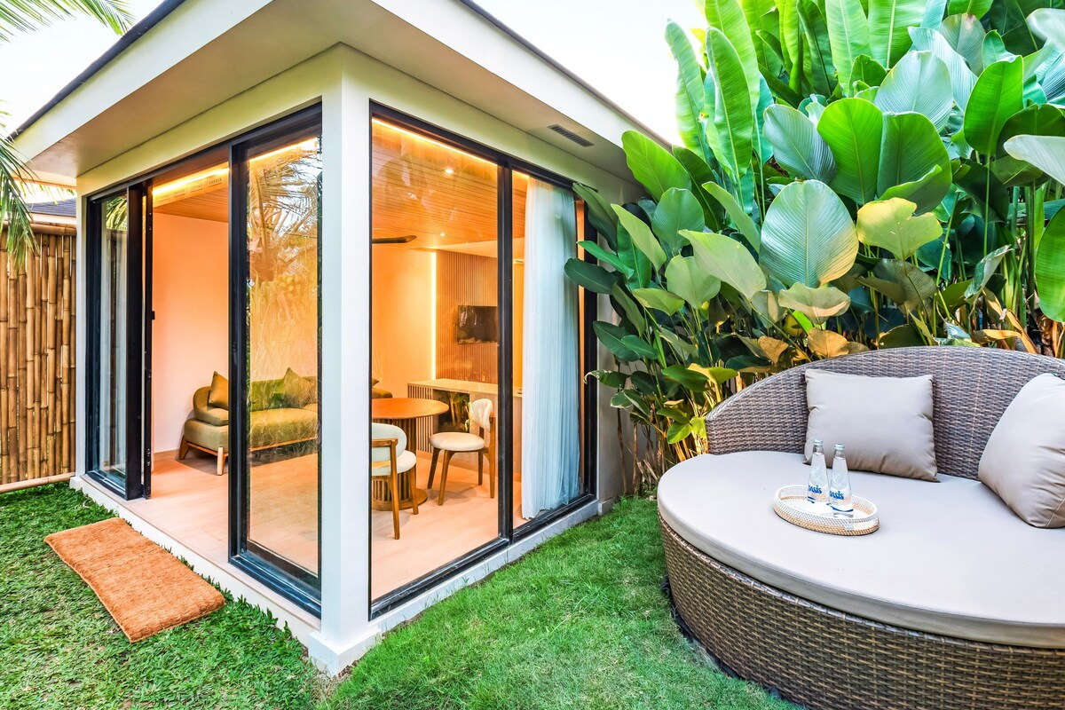 Una casa moderna con un patio cubierto de vidrio, un sofá redondo de mimbre y abundante vegetación.