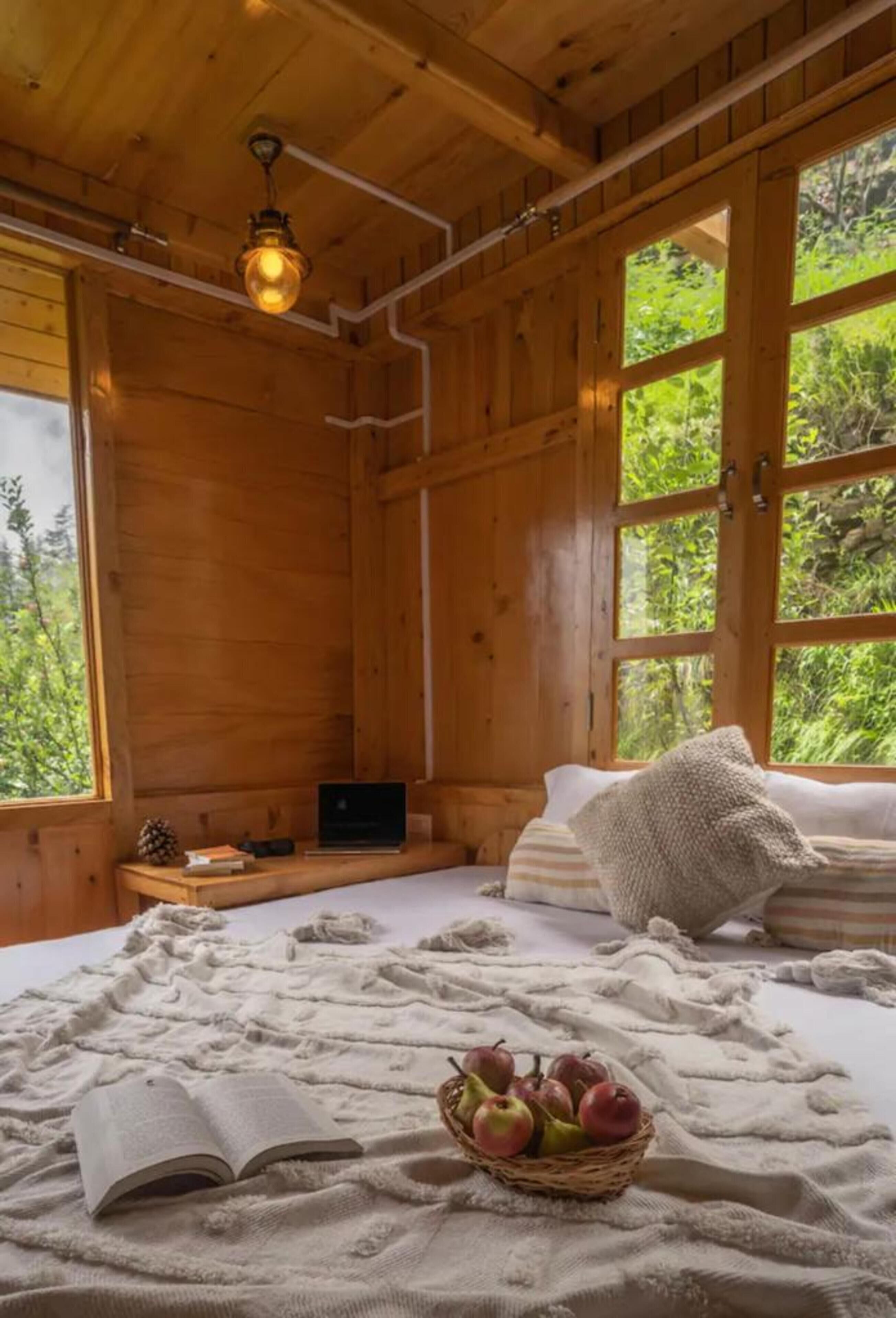 A wooden cabin interior with a bed, a laptop, a book, and a basket of fruit.