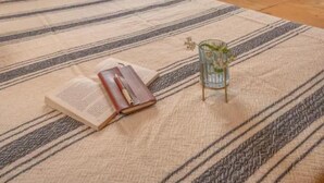Egy hangulatos, fából készült szoba, ferde mennyezettel, nagy ablakkal, csíkos szőnyeggel, és egy kis asztallal, melyen egy könyv és egy üveg váza található.