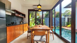 A modern kitchen with wooden cabinets, a dining area with a table and chairs, and a view of a swimming pool through large windows. - Villa Movida Canggu by Nagisa Bali (Kerobokan)