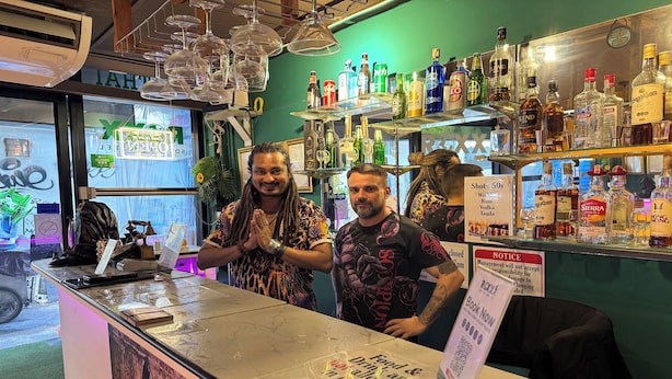 Un espace bar avec des étagères remplies de bouteilles d’alcool, un comptoir avec un menu et un panneau indiquant les différents types de boissons et leurs prix.