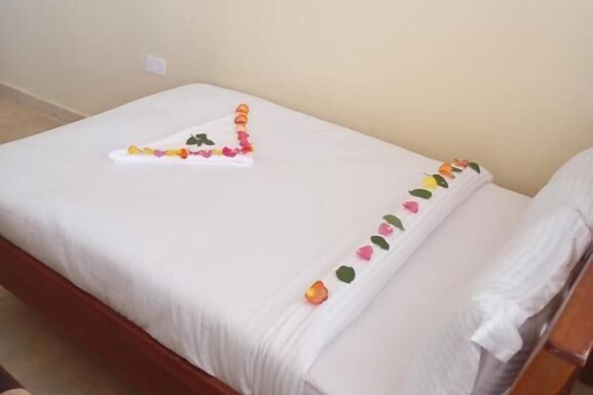 A single bed with a white sheet, a small decorative item on the bed, and a plain wall with a light switch.