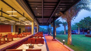 A restaurant with a long counter and multiple tables, a covered outdoor seating area, and a view of palm trees and greenery. - Zo Selections Gokarna by Zostel (Gokarna)