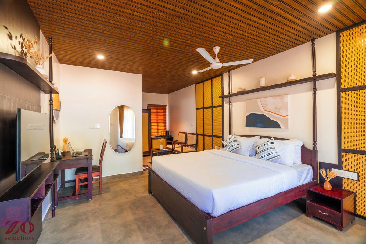 Une chambre à coucher comprenant un lit, un bureau, une chaise, un téléviseur, un ventilateur de plafond et une fenêtre avec des stores.