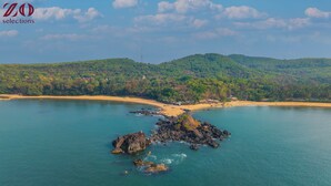 An aerial view of a coastal area with a small rocky island surrounded by clear blue water, a sandy beach, and a forested hill in the background. - Zo Selections Gokarna by Zostel (Gokarna)