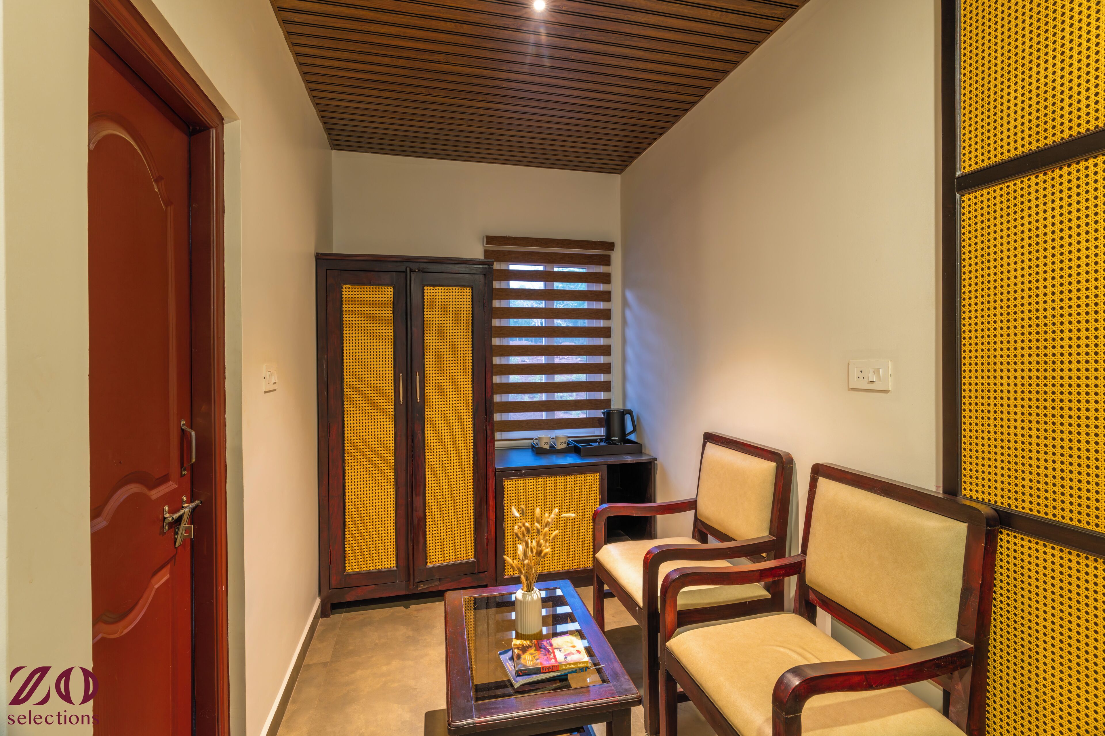 Una habitación con techo de madera, una puerta, un área de descanso con sillas, una mesa con una revista y una ventana con persianas.