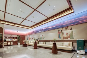 Un hall d’entrée spacieux avec un plafond haut, des luminaires encastrés, un coin salon avec des canapés, un espace dédié au café et une grande fresque murale.