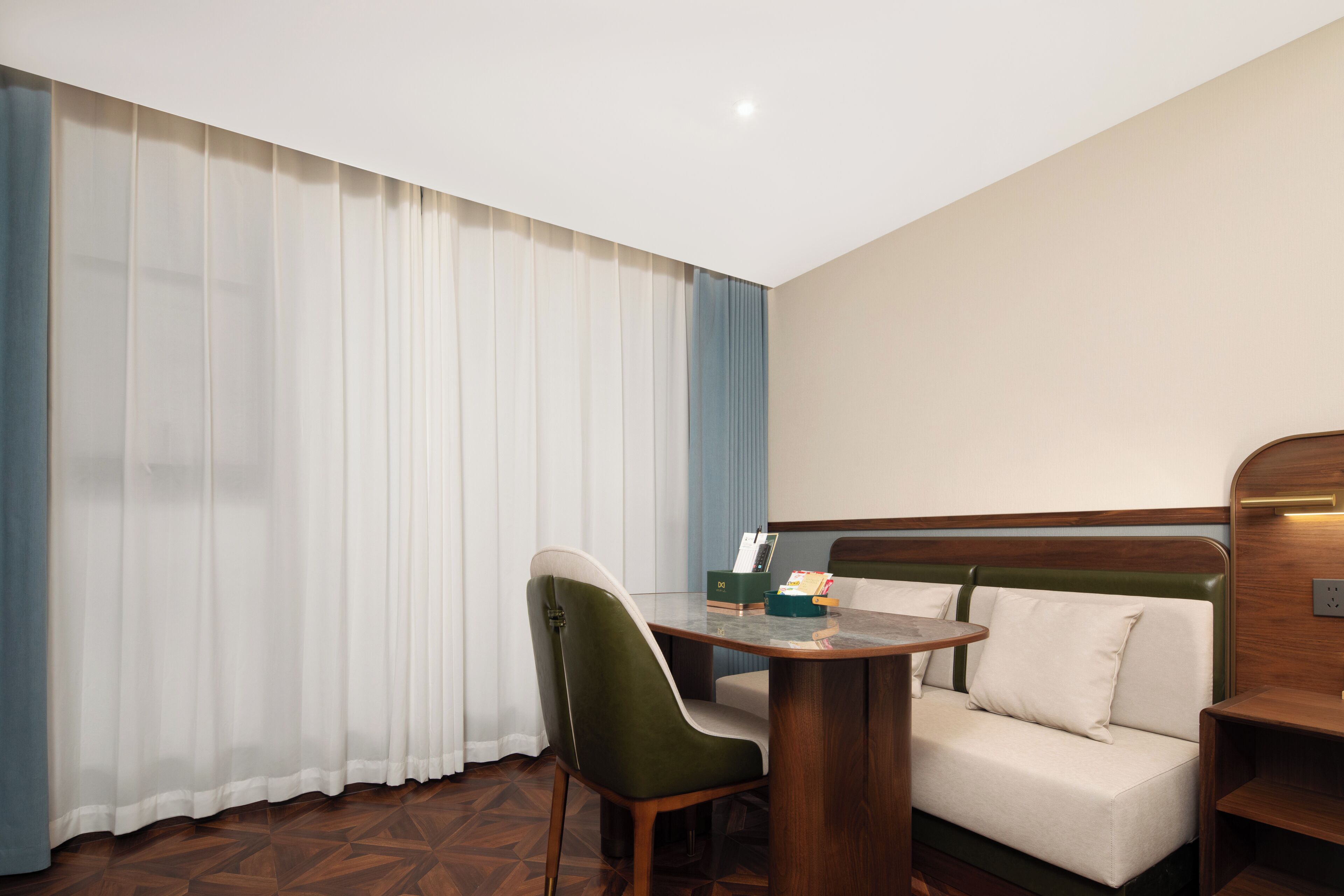 Quarto de hotel com cama, uma cadeira verde, uma mesinha e um criado-mudo.