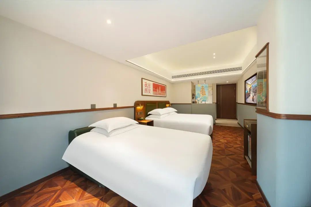 A hotel room with two beds, a wooden floor, a wall-mounted TV, and a framed picture.