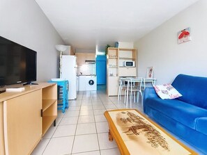 Ein Wohnzimmer mit Fernseher, blauem Sofa, Couchtisch und einer Küche im Hintergrund.