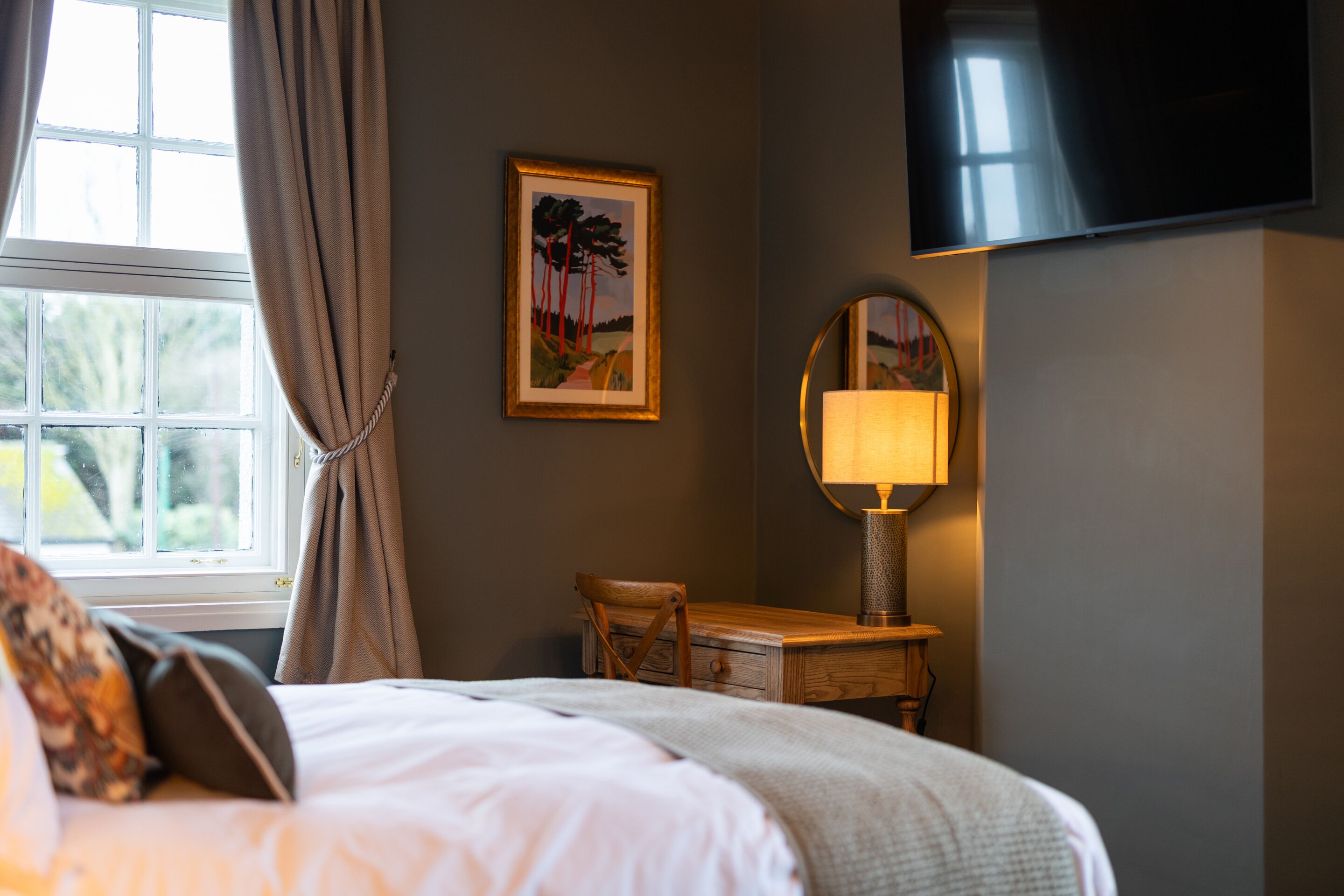 Ein Hotelzimmer mit Bett, Schreibtisch mit Lampe, Fernseher und Fenster mit Vorhängen.