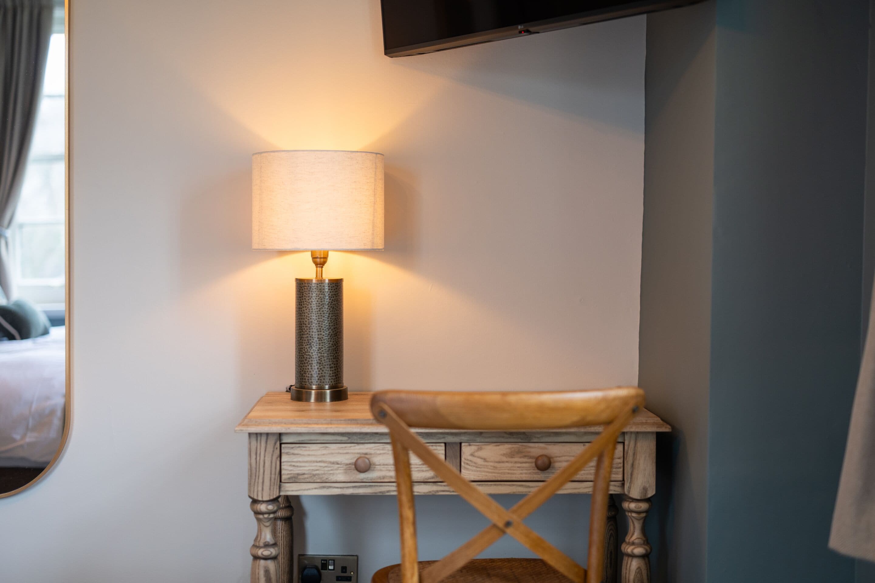 A wooden bedside table with a lamp, a chair, and a flat-screen TV mounted on the wall.