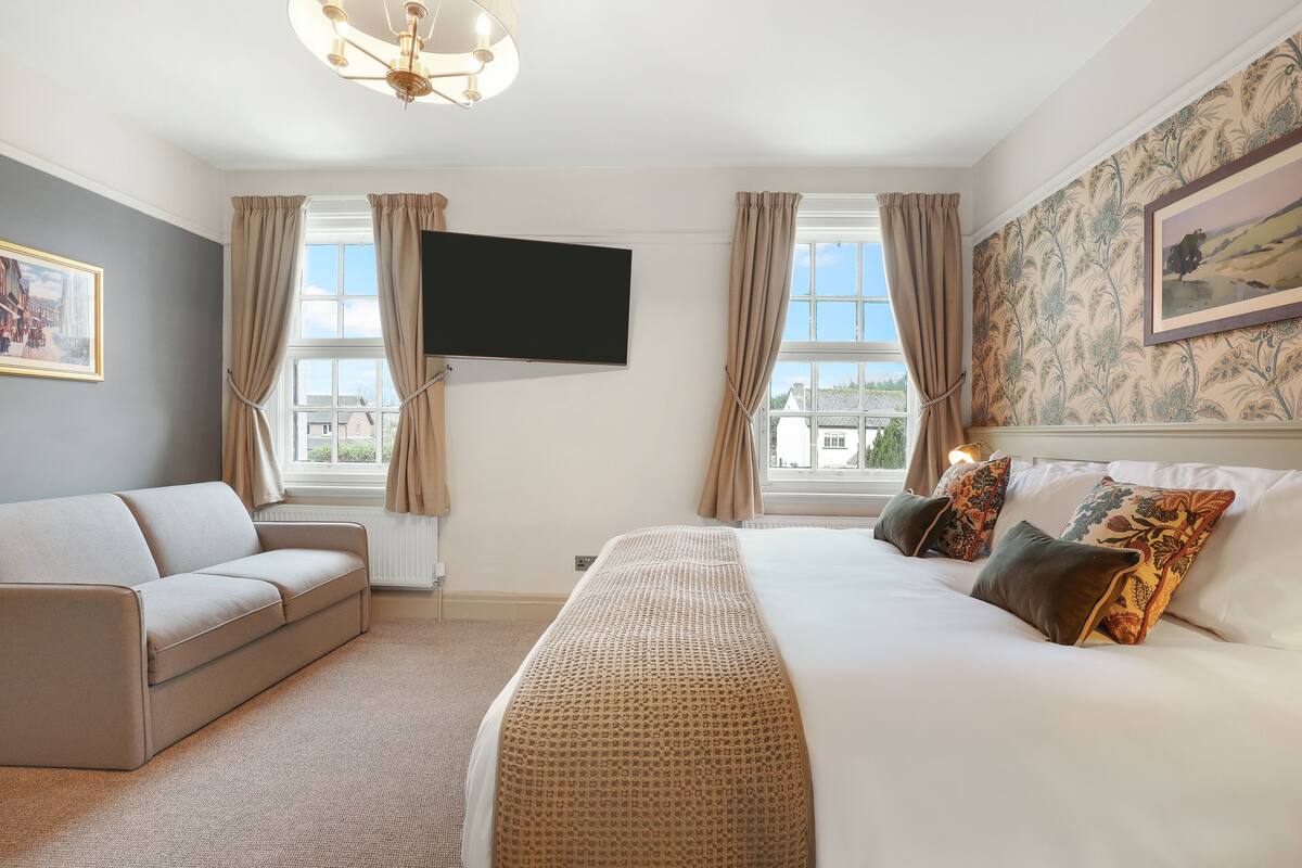 Una habitación de hotel con cama, sofá, televisión y ventana con cortinas.