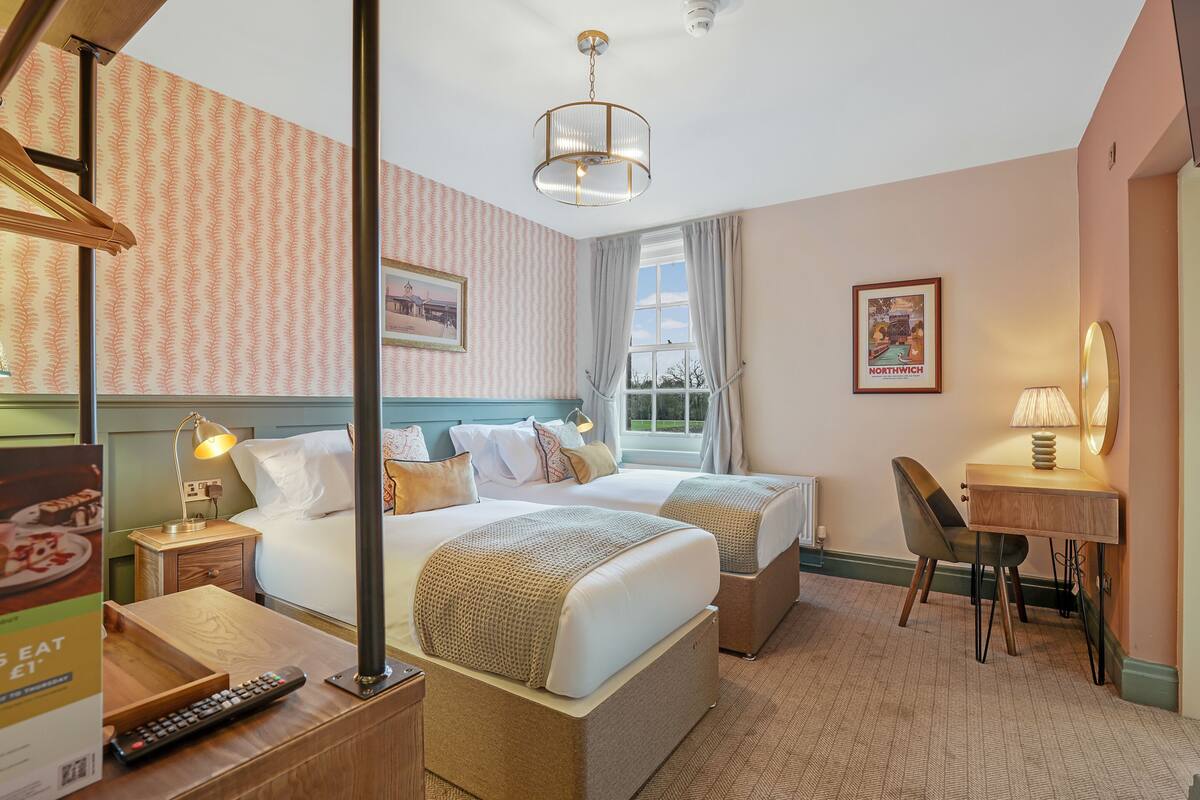 Habitación de hotel con cama, dos mesitas de noche, escritorio, silla, lámpara y ventana con cortinas.