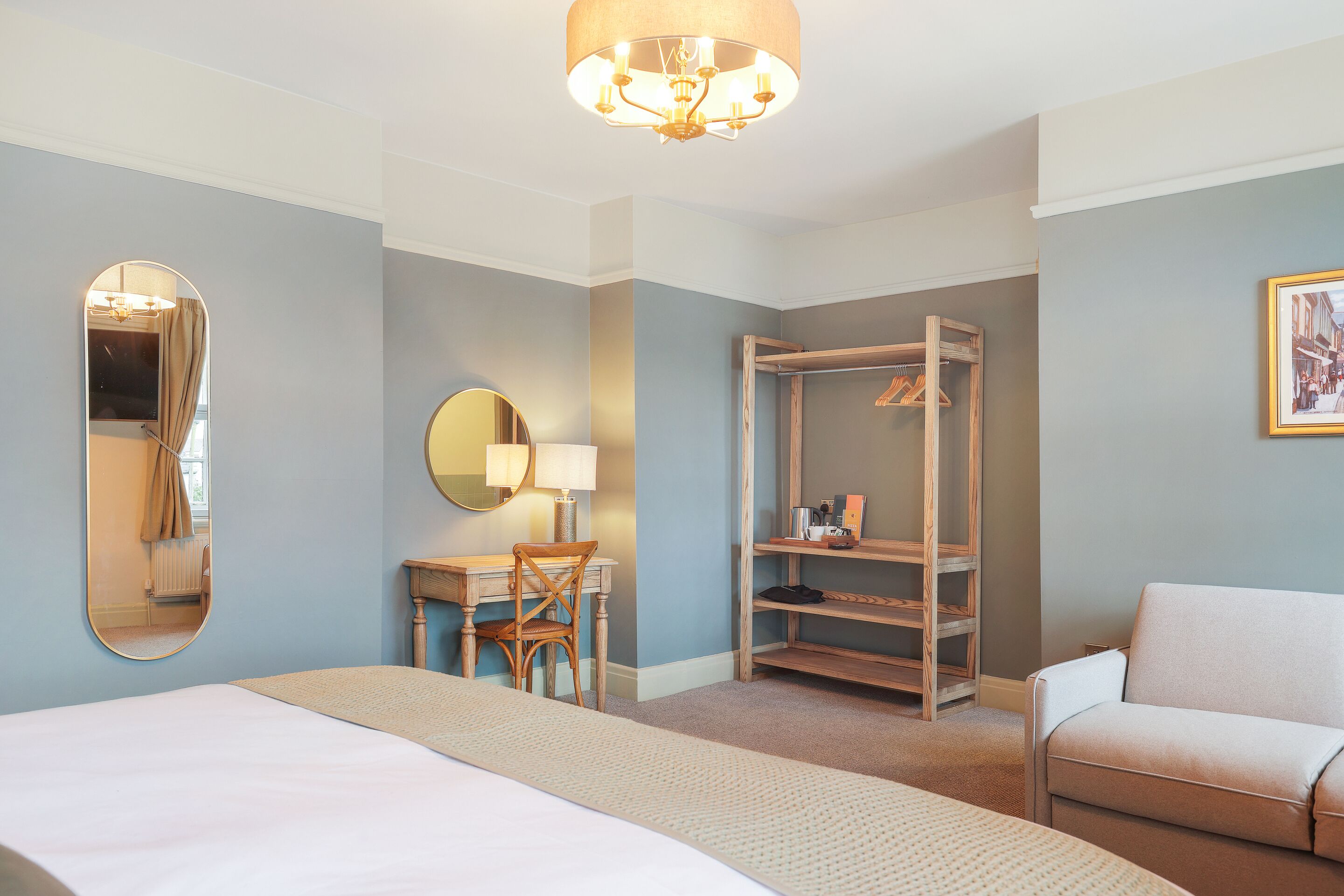 A hotel room with a bed, a desk with a chair, a wardrobe, a sofa, a mirror, and a chandelier.