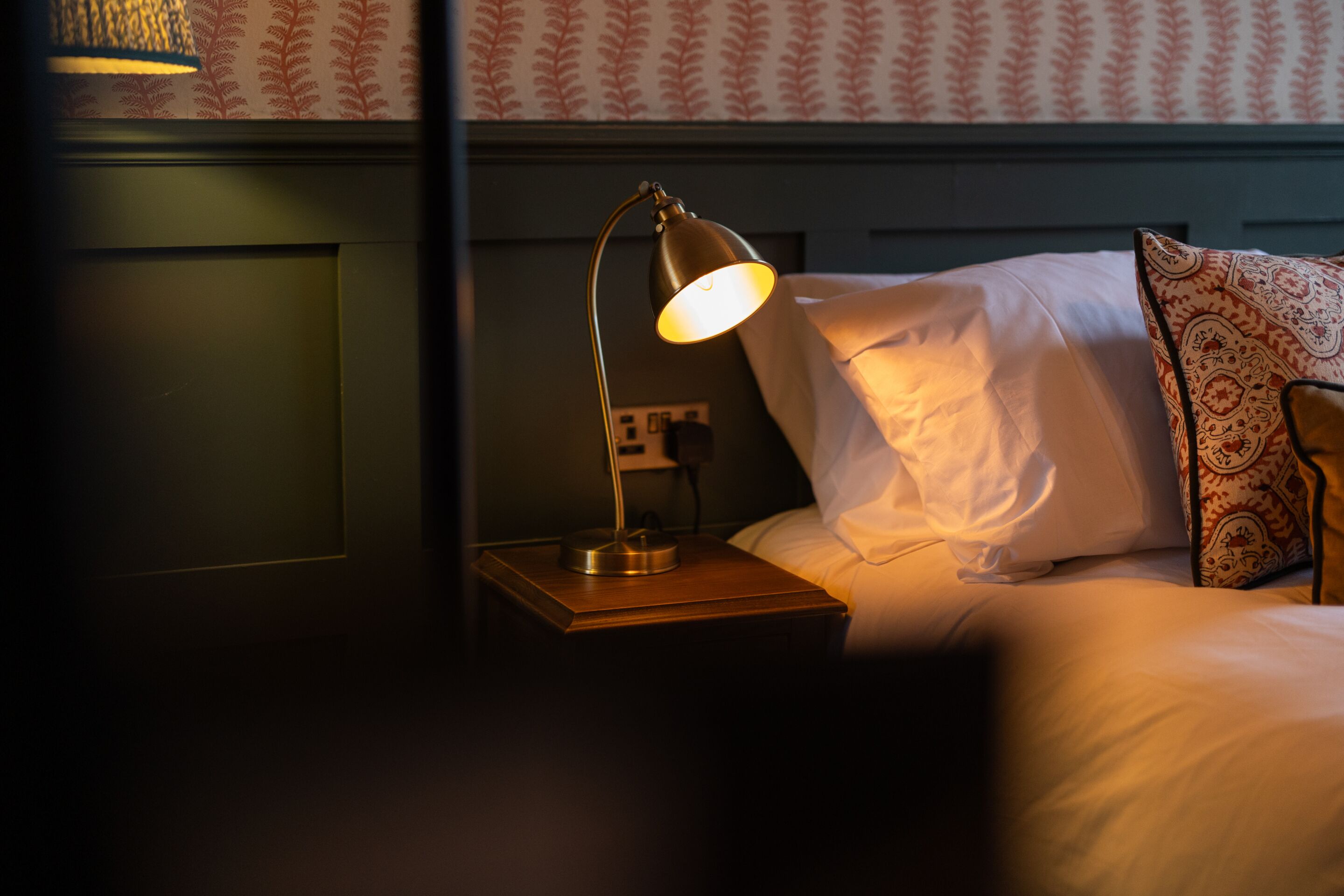 A neatly made bed with a patterned pillow, a bedside lamp, and a wall with a floral wallpaper pattern.