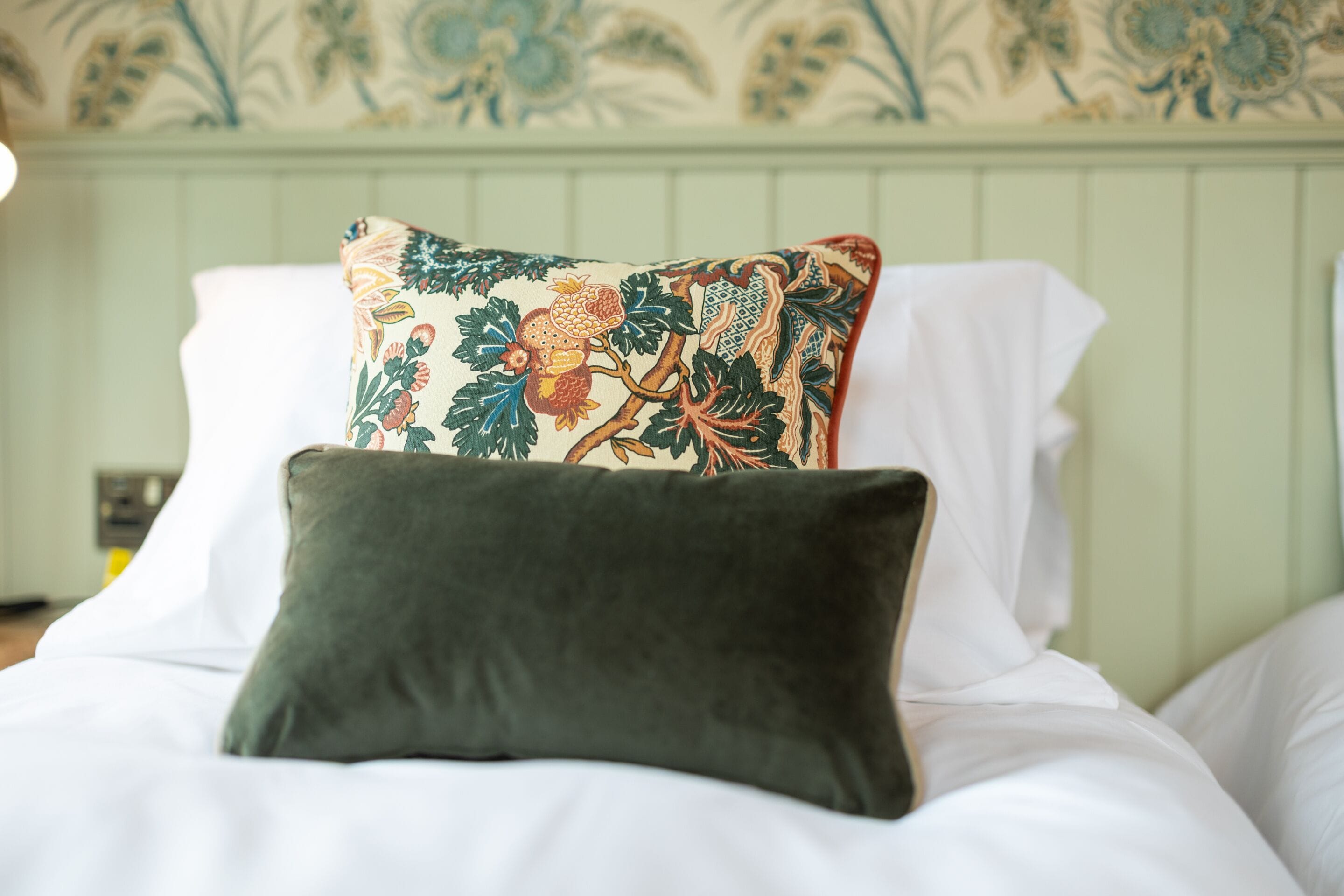 Un lit avec un coussin décoratif et un coussin uni, un mur vert recouvert d’un papier peint à motifs floraux et une tête de lit blanche.