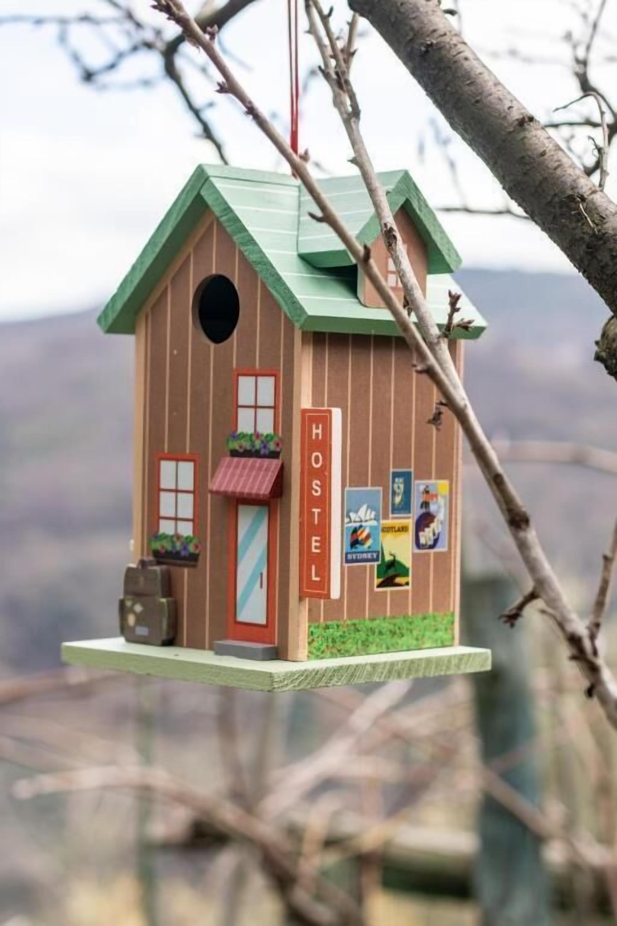 Un comedero de pájaros con techo verde, puerta roja y un letrero que dice 'HOSTEL'.