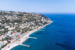 A coastal town with buildings along the beachfront, a clear blue sea, and a hilly backdrop. - The Big Blue (Ospedaletti)