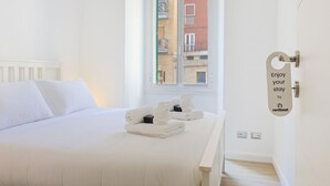 A white bed with white pillows, a window with a view of buildings, and a door with a sign saying 'Enjoy your stay by rentbeat'. - Tuscolana Costantino's Apartment by Rentbeat (Roma)