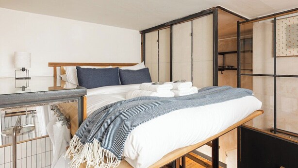A bed with a wooden frame, a white mattress, and a blue and white striped blanket. - New! Bright Brixton 1BR | Walk to Market & Tube (Greater London)